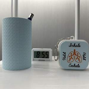 May include: A light blue cylindrical pen holder with a textured grid pattern, a small white digital clock displaying 8:55, and a square light blue keychain featuring a graphic of lungs with the text Inhale, Hold, Hold, and Exhale.