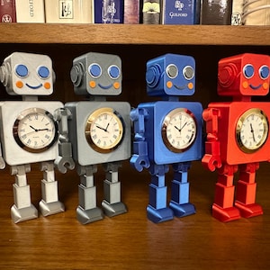 May include: Four robot-shaped clocks in silver, gray, blue, and red. Each robot has a square body with a clock face, arms, legs, and a head with eyes and a smile. The clocks are on a wooden surface.