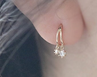 May include: Gold hoop earrings with a small, dangling, clear gemstone. The earrings are worn on an ear, with the brand name "ARTEAFINITY" visible at the bottom left. The earrings are a delicate piece of jewelry.