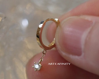 May include: A gold hoop earring with a small diamond dangling from it. The earring is held in a hand.