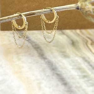 May include: A pair of gold-colored hoop earrings with dangling chain accents. The earrings are displayed on a thin, gold-colored rod against a blurred background. The hoops have a simple, elegant design.