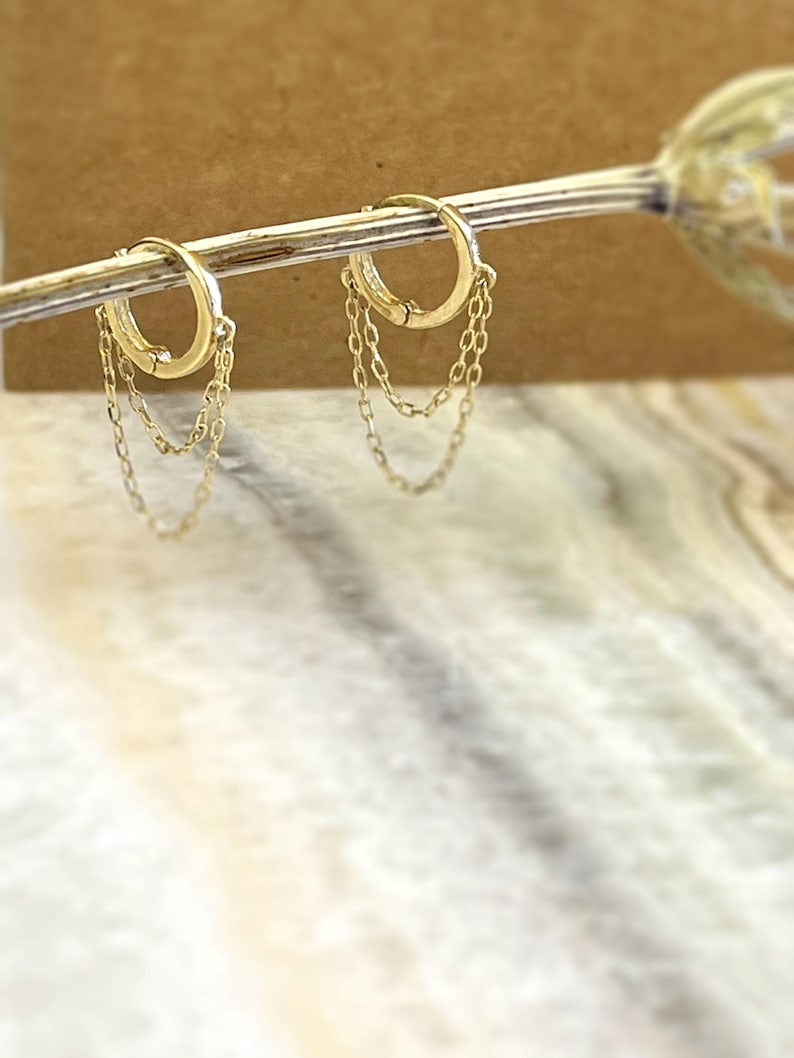 May include: A pair of gold-colored hoop earrings with dangling chain accents. The earrings are displayed on a thin, gold-colored rod against a blurred background. The hoops have a simple, elegant design.