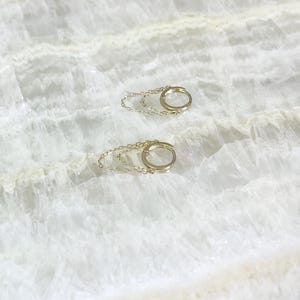 May include: A pair of gold-colored hoop earrings with a delicate chain detail. The earrings are small and circular, with a simple, elegant design. The background is a white, marble-like surface.