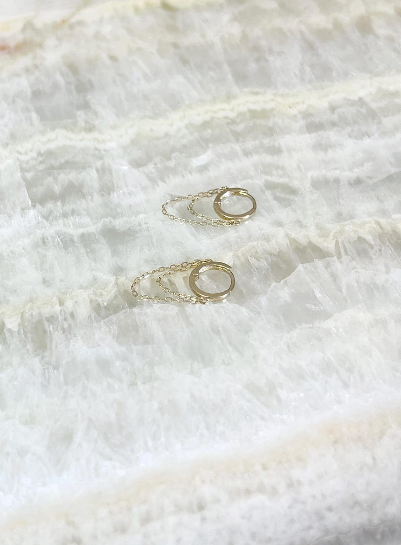 May include: A pair of gold-colored hoop earrings with a delicate chain detail. The earrings are small and circular, with a simple, elegant design. The background is a white, marble-like surface.