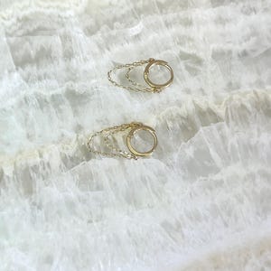 May include: Two gold-colored hoop earrings with a delicate chain detail. The earrings are circular with a small clasp and a chain that dangles below. The background is a white, marbled surface.