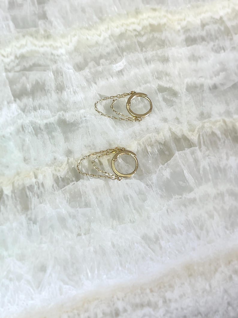 May include: Two gold-colored hoop earrings with a delicate chain detail. The earrings are circular with a small clasp and a chain that dangles below. The background is a white, marbled surface.