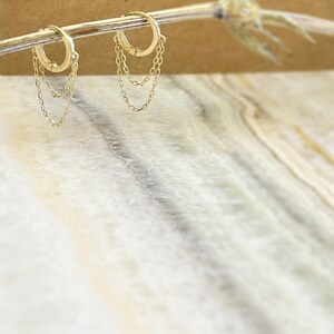 May include: A pair of gold hoop earrings with delicate chain dangles. The earrings are displayed on a thin, light-colored branch against a blurred background. The hoops are round and have a simple, elegant design, perfect for everyday wear.