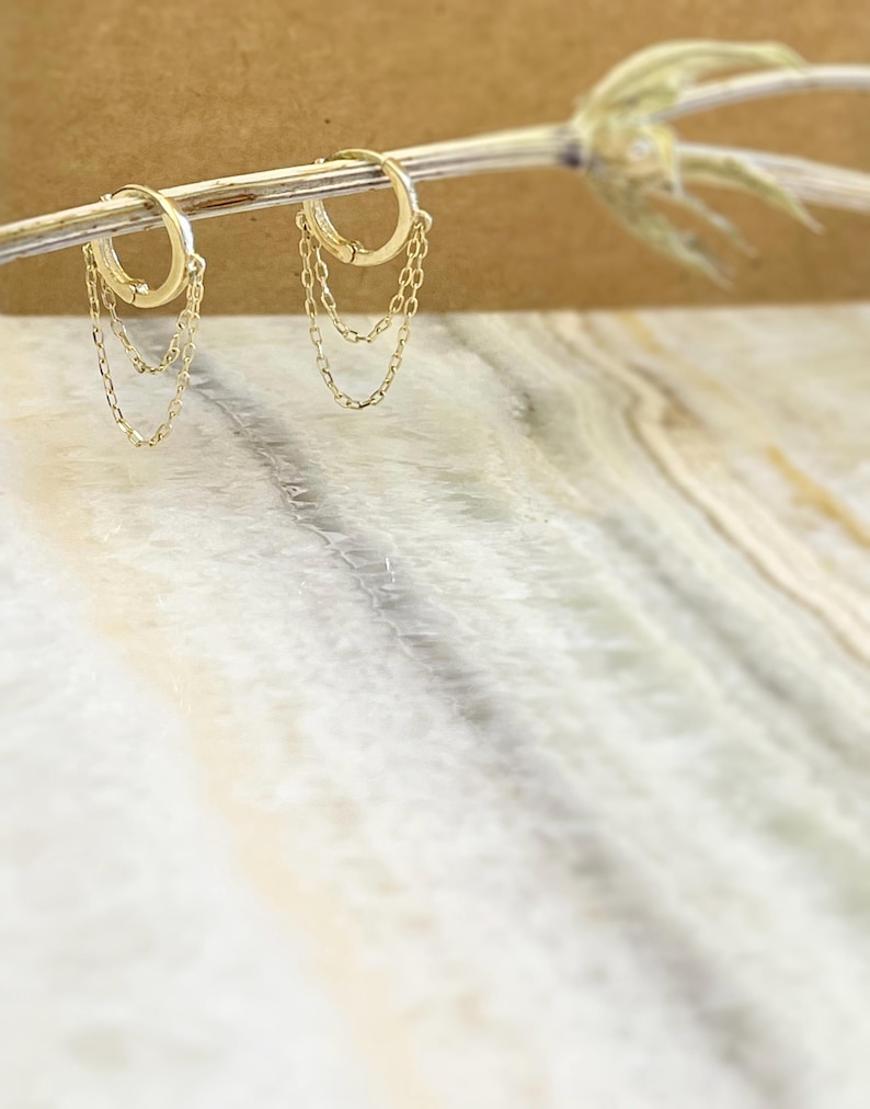 May include: A pair of gold hoop earrings with delicate chain dangles. The earrings are displayed on a thin, light-colored branch against a blurred background. The hoops are round and have a simple, elegant design, perfect for everyday wear.