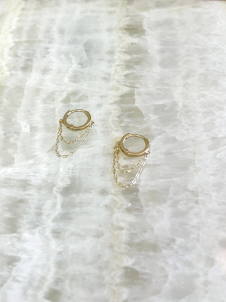 May include: A pair of gold-colored hoop earrings with delicate chain dangles. The earrings are displayed on a white, marbled surface, showcasing their simple yet elegant design. The chains hang gracefully from the hoops, adding a touch of movement.