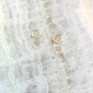 May include: A pair of gold-colored hoop earrings with three delicate chain dangles. The earrings are displayed on a white, marbled surface, creating a minimalist and elegant aesthetic. The chains hang gracefully from the hoops.