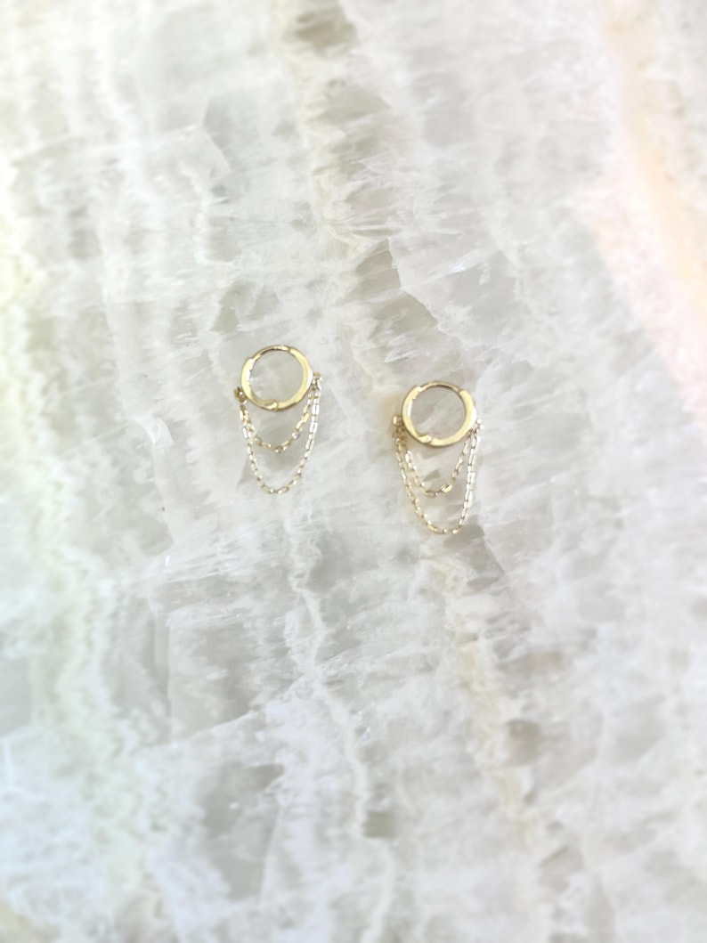 May include: A pair of gold-colored hoop earrings with three delicate chain dangles. The earrings are displayed on a white, marbled surface, creating a minimalist and elegant aesthetic. The chains hang gracefully from the hoops.