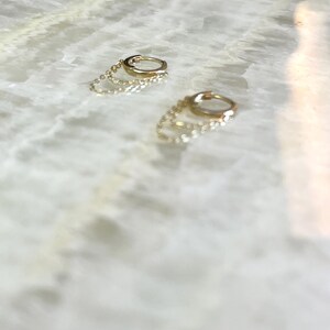 May include: Two gold hoop earrings with delicate chain accents. The earrings are small and circular, with a polished finish. The chains hang from the hoops, adding a subtle dangle. The earrings are set against a light-colored, marble-like surface.