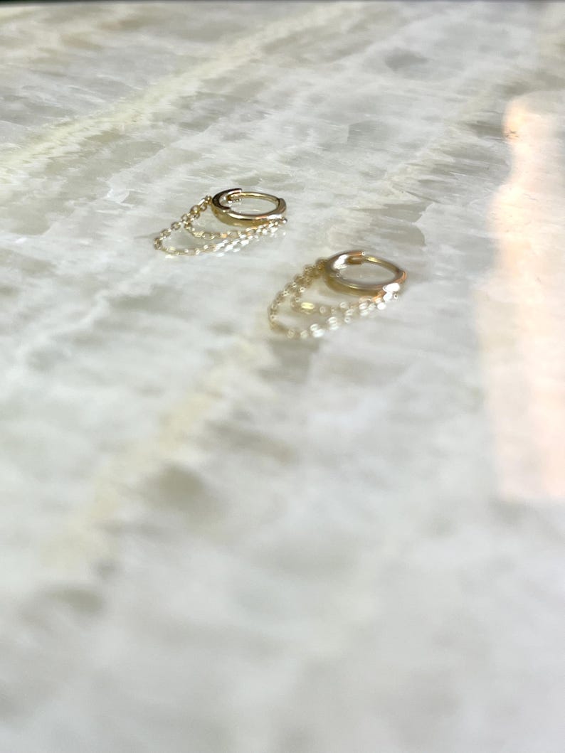 May include: Two gold hoop earrings with delicate chain accents. The earrings are small and circular, with a polished finish. The chains hang from the hoops, adding a subtle dangle. The earrings are set against a light-colored, marble-like surface.