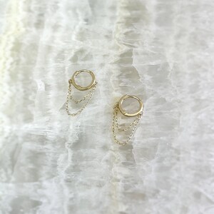 May include: A pair of gold-colored hoop earrings with delicate chain dangles. The earrings are displayed on a white, marbled surface, creating a simple yet elegant presentation. The chains hang gracefully from the hoops, adding movement and visual interest.