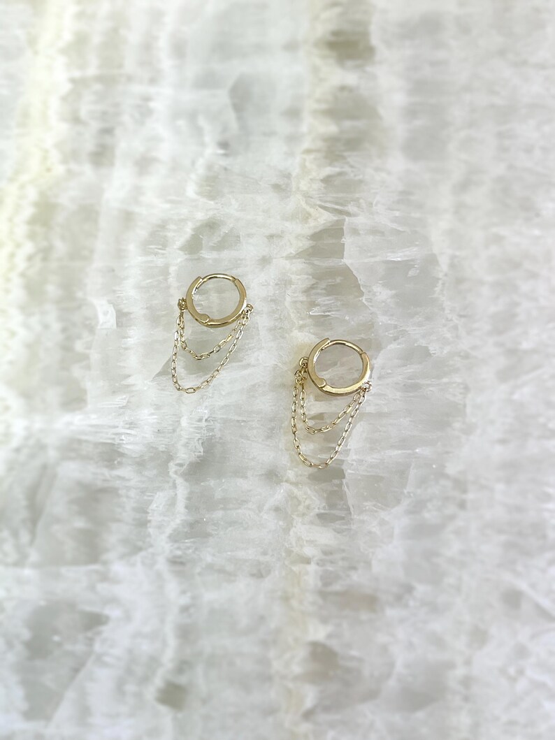 May include: A pair of gold-colored hoop earrings with delicate chain dangles. The earrings are displayed on a white, marbled surface, creating a simple yet elegant presentation. The chains hang gracefully from the hoops, adding movement and visual interest.