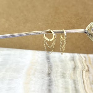 May include: A pair of gold-colored hoop earrings with delicate chain dangles. The earrings are displayed on a light-colored surface with a blurred background. The chains hang gracefully from the hoops, adding a touch of elegance.