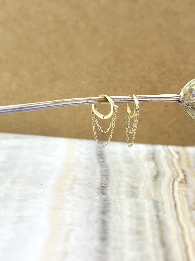 May include: A pair of gold-colored hoop earrings with delicate chain dangles. The earrings are displayed on a light-colored surface with a blurred background. The chains hang gracefully from the hoops, adding a touch of elegance.
