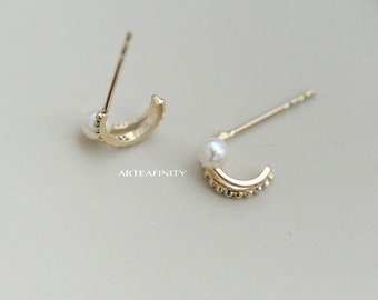 May include: A pair of gold-colored crescent-shaped stud earrings. Each earring features a small, round pearl at the top and a row of tiny beads along the curved edge. The earrings are displayed on a neutral background.