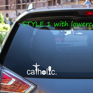 May include: A white vinyl sticker on the back window of a silver car. The sticker says "catholic." with a cross and a chalice.