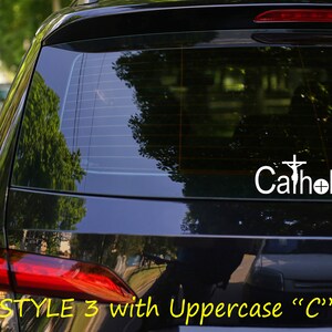 May include: A black car with a white vinyl decal on the rear window. The decal says "Catholic" with a cross and a monstrance.