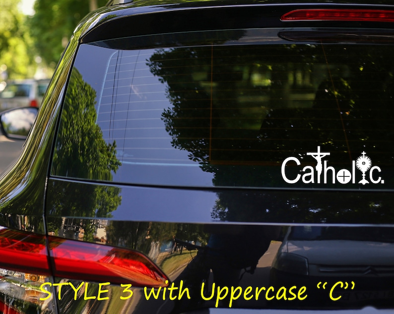 May include: A black car with a white vinyl decal on the rear window. The decal says "Catholic" with a cross and a monstrance.