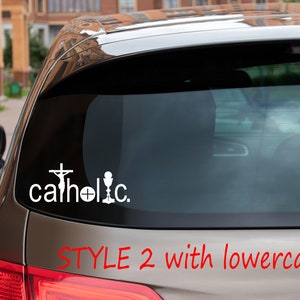 May include: A silver car with a white vinyl decal on the back window that reads "catholic" with a cross, chalice, and host. The text "STYLE 2 with lowercase "c"" is written in red below the decal.