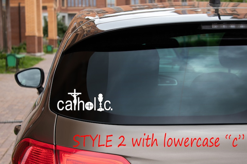 May include: A silver car with a white vinyl decal on the back window that reads "catholic" with a cross, chalice, and host. The text "STYLE 2 with lowercase "c"" is written in red below the decal.
