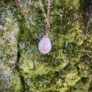 Puede incluir: Un delicado collar de cadena dorada con un colgante blanco en forma de l&aacute;grima con detalles de hoja de oro. El colgante cuelga sobre un fondo verde musgoso.