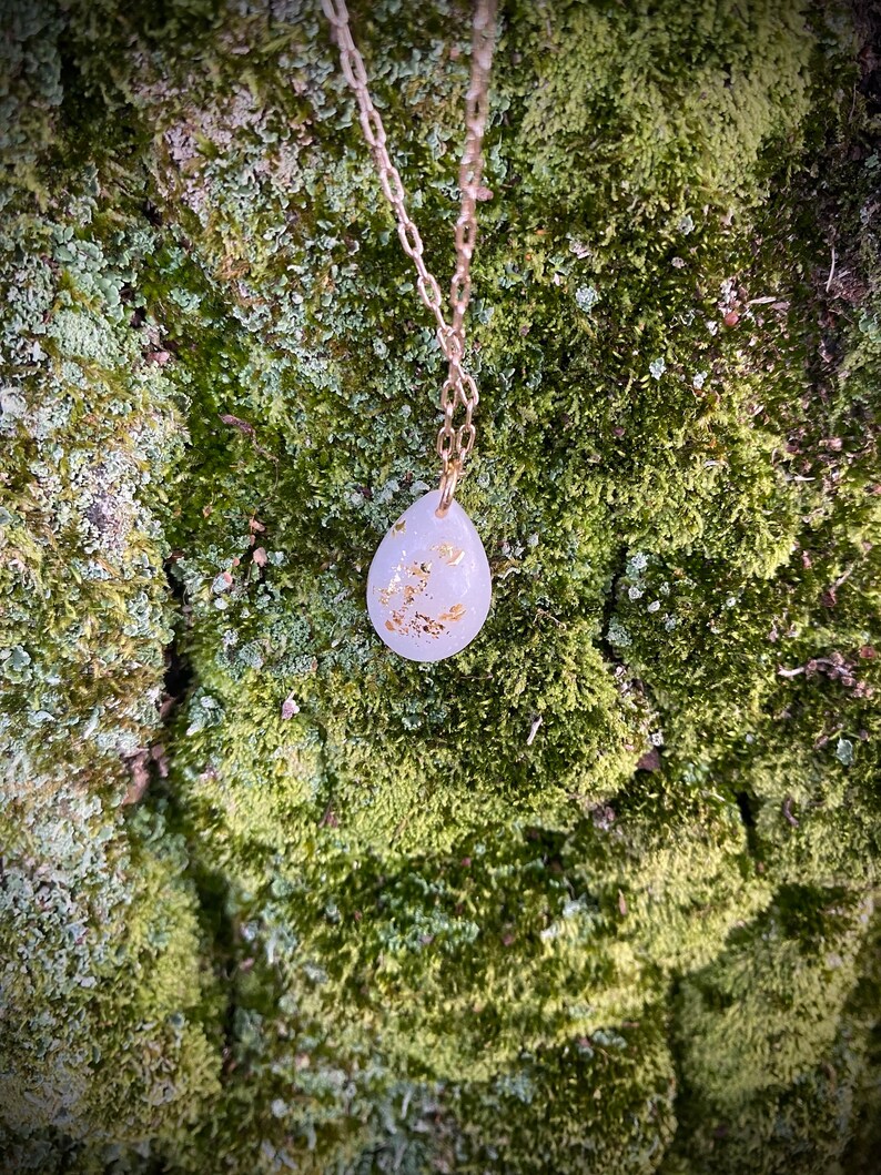 Puede incluir: Un delicado collar de cadena dorada con un colgante blanco en forma de l&aacute;grima con detalles de hoja de oro. El colgante cuelga sobre un fondo verde musgoso.