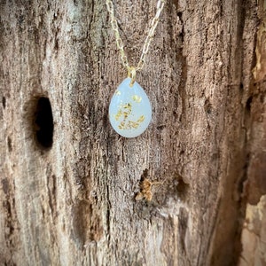 Puede incluir: Un collar de cadena dorada con un colgante blanco en forma de l&aacute;grima con motas doradas. El colgante cuelga sobre un fondo de madera.