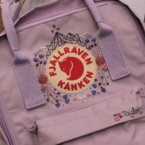 Puede incluir: Una mochila Fjällräven Kånken de color morado con un diseño de bordado floral alrededor del logotipo. La mochila tiene un pequeño bolsillo en la parte delantera con el nombre "Taylor" bordado.