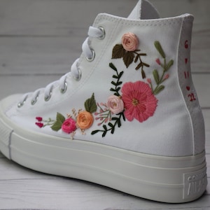 May include: White high-top sneakers with embroidered pink and peach flowers and green leaves. The date "6.11.22" is embroidered on the side of the shoe.
