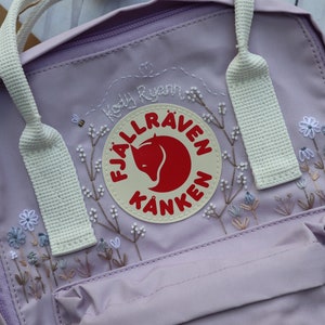 Puede incluir: Una mochila Fjällräven Kånken de color lavanda con correas blancas y un diseño floral bordado en blanco. El logotipo es visible con un zorro rojo en un círculo crema con el texto "Fjällräven Kånken". La mochila está personalizada con el texto "Kody Ryan".