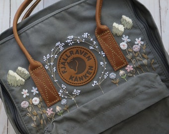 Puede incluir: Una mochila Fjällräven Kånken gris con una correa de cuero marrón y flores y hojas blancas bordadas. La mochila tiene una cremallera en el lateral y un pequeño bolsillo en la parte delantera.