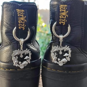 May include: Two black leather boots with silver heart-shaped charms. Each charm has a mushroom and snail design. The boots have a black pull tab with gold lettering.