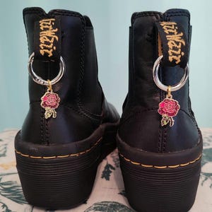 May include: Black leather ankle boots with a gold chain and rose charm. The boots have a thick platform sole and a pull-on design. The text "Dr. Martens" is printed on the side of the boots.