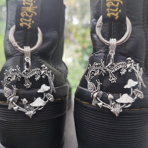 May include: Black leather boots with silver mushroom and snail charms hanging from the pull loops. The charms are detailed with fungi and foliage. The boots have chunky soles, set against a blurred backdrop.