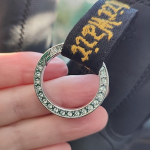 May include: A silver key chain with a round, clear crystal-encrusted ring. The ring is attached to a black strap with the word "Dr. Martens" embroidered in yellow.
