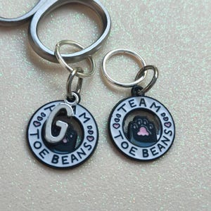 May include: Two black and white enamel charms with silver jump rings. The charms are round and have a black paw print inside a white circle. The text "Team Toe Beans" is written around the outside of the circle. One charm has the letter "G" in the center of the circle.