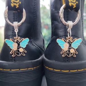 May include: Two black leather boots with yellow stitching, each featuring a silver charm. The charms are bee-shaped, with gold-colored bodies, light blue wings, and white flower details. The boots are shown from the back.