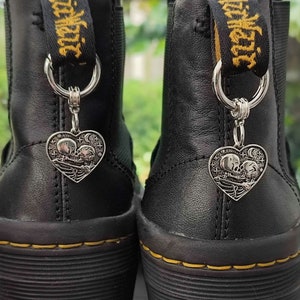 May include: A pair of black leather ankle boots featuring yellow stitching and black pull tabs with yellow text. Each boot is adorned with a silver-toned heart-shaped charm depicting two skeletons embracing under a starry night sky.