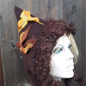 Puede incluir: Un gorro de fieltro marrón con capucha y ribete de piel rizada marrón. El gorro tiene una parte superior puntiaguda con dos hojas de fieltro en tonos naranja y amarillo.