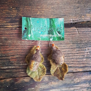 Puede incluir: Un par de pendientes de fieltro en forma de hoja en tonos marrones y verdes. Los pendientes presentan una pequeña bellota de fieltro marrón con una cuenta marrón en la parte superior. Los pendientes cuelgan de un gancho plateado.