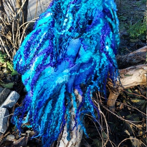 Puede incluir: Una bufanda de lana azul y morada con largos mechones rizados. La bufanda está drapeada sobre una rama de árbol.
