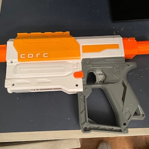 May include: A white and orange toy gun with a gray grip. The gun has a black barrel and the word "CORC" printed on the side.