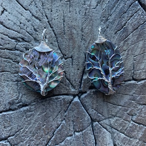 May include: Two abalone shell pendants with silver wire tree of life designs. The iridescent shells are round and oval, with the wire wrapped around the edges and forming the tree's branches. The pendants are displayed on a textured, gray surface.