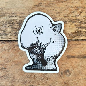 Può includere: Un adesivo bianco con un'illustrazione in bianco e nero di una creatura dei cartoni animati. La creatura ha una testa grande, un orecchio piccolo e un corpo arrotondato. L'adesivo è su una superficie di legno.