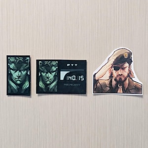 May include: Three stickers featuring characters from a video game. One sticker shows a character's face in a green and black color scheme. Another sticker shows the same character's face with a digital display. The third sticker shows a character saluting.