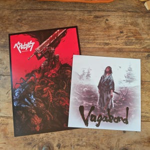 May include: Two art prints on a wooden surface. One print features a warrior with a large sword against a red background with Japanese text. The other print shows a samurai with the word "Vagabond" in black script.