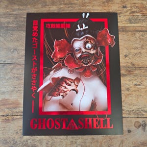 Può includere: Un poster nero con un bordo rosso con un'illustrazione di cyborg di "Ghost in the Shell". L'opera raffigura una figura femminile con componenti meccanici esposti e testo giapponese. Il titolo "Ghost in the Shell" è stampato in rosso.